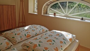 Ein Schlafzimmer mit einem Bett, einem Holzschrank, einer Hockerleiter, einem Fenster mit Blick auf Grün und einem kleinen Regal mit einer Kerze.