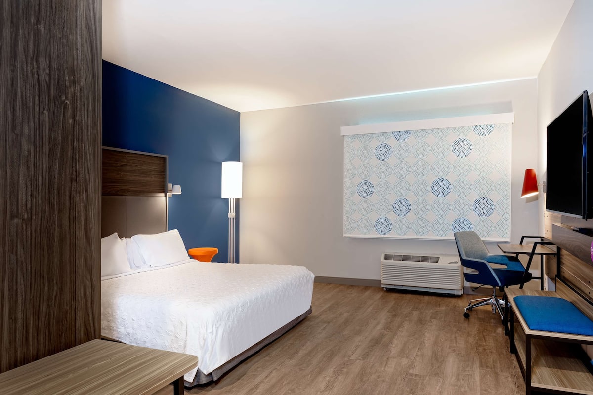 Habitación de hotel con cama, escritorio, silla, televisión y ventana con cortina estampada.
