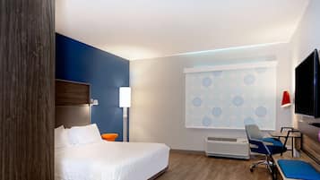 A hotel room with a bed, a desk, a chair, a TV, and a window with a patterned curtain.
