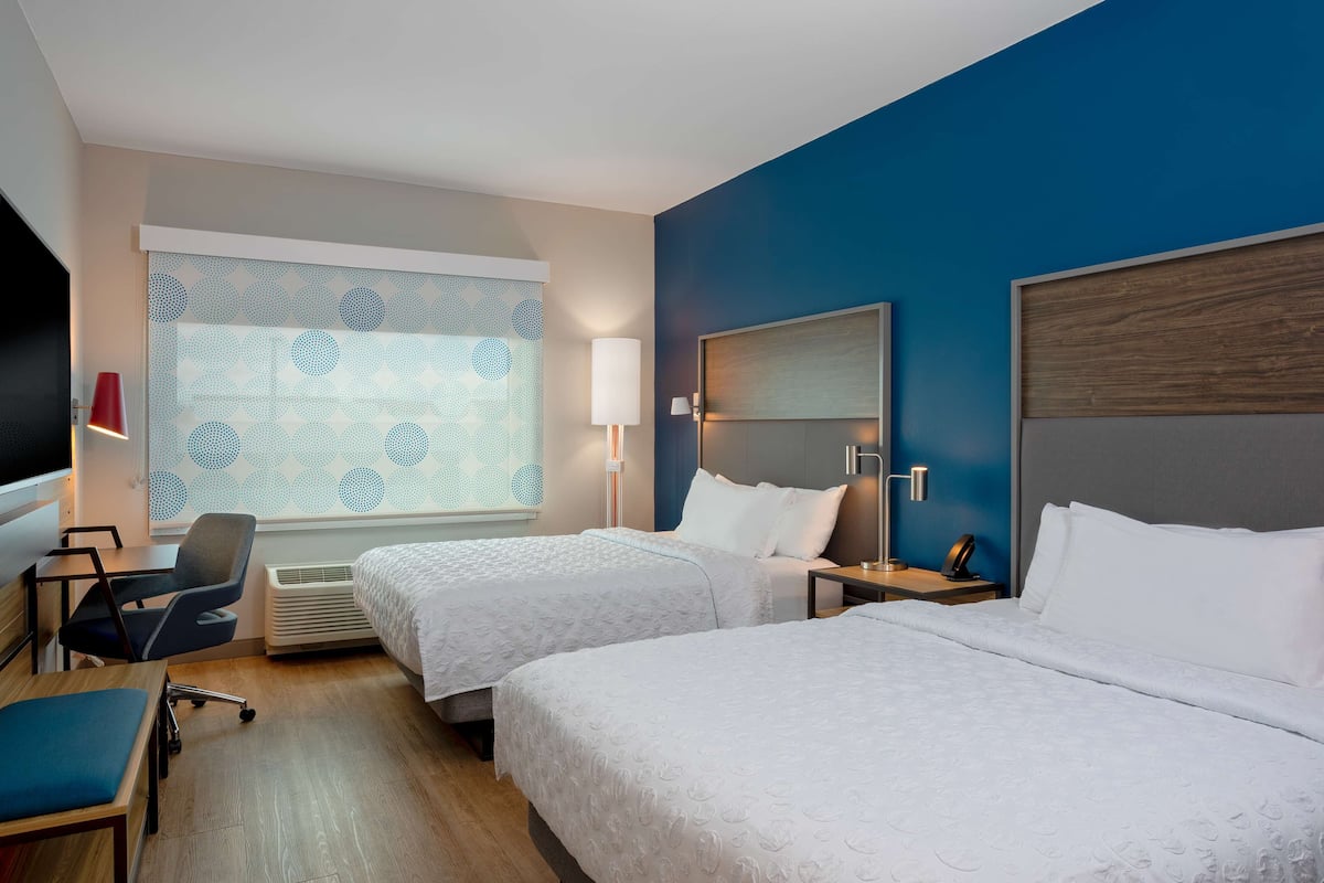 Habitación de hotel con dos camas, un escritorio, una silla, un ventanal con cortinas estampadas y una pared de acento azul.