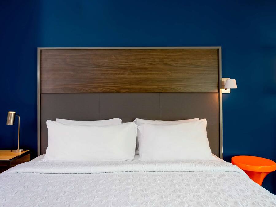 A neatly made bed with white linens and a wooden headboard, a bedside table with a lamp, and an orange chair in a room with blue walls.