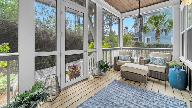 Une terrasse extérieure couverte, aménagée avec des meubles en osier, des plantes en pot et offrant une vue sur une maison et des arbres.