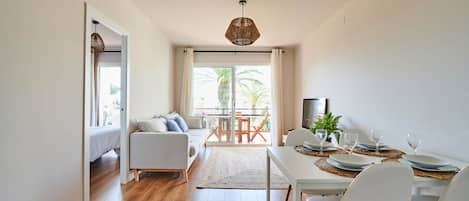A modern living room with a sofa, dining table, and chairs. There is a balcony with a view of palm trees.