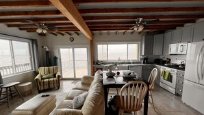A cozy living room with a sofa, chairs, and a dining table. There is a kitchen with a stove, microwave, and cabinets. A large window offers a view of the ocean.