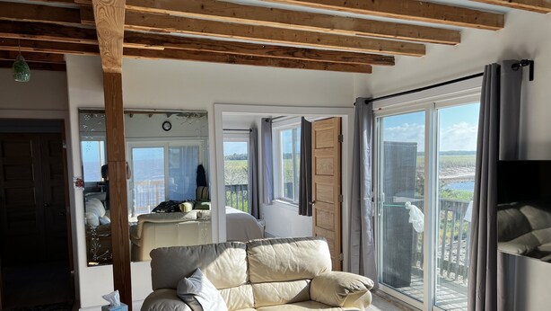 A spacious living room with a large beige sofa, a wooden ceiling, and a sliding glass door leading to a balcony with a view of the countryside.