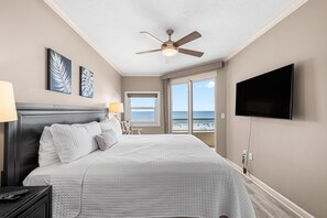 A hotel room with a bed, a television, a ceiling fan, artwork on the wall, and a window with a view of the beach.
