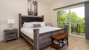 A modern bedroom with a large bed, a nightstand, a wooden bench, and a balcony with a view of greenery.