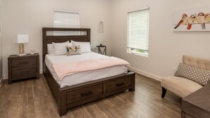 A bedroom with a wooden bed, bedside tables, a nightstand, a chair, and two windows with blinds.