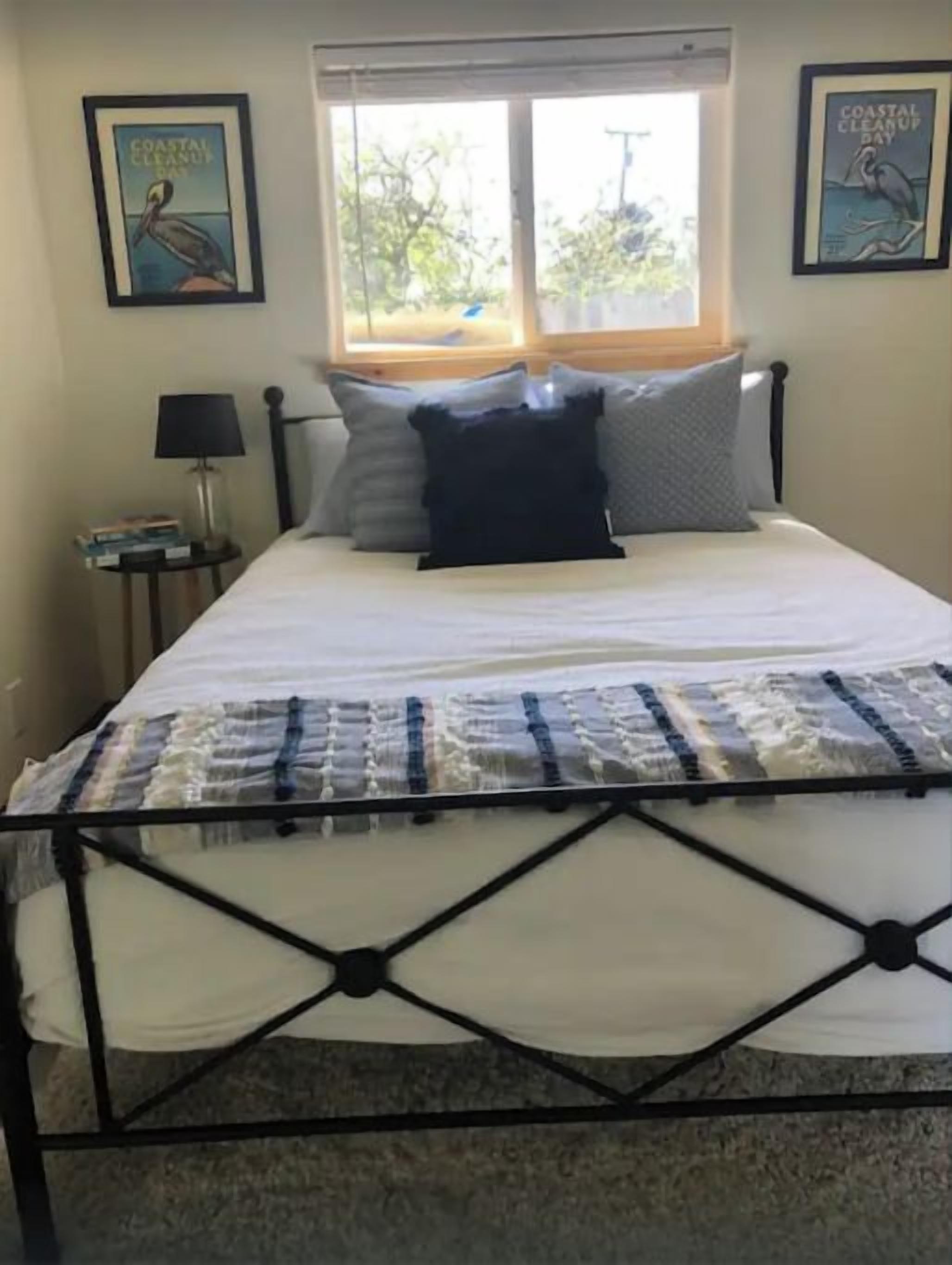 Una cama con estructura metálica, una ventana con persianas, y dos cuadros en la pared.