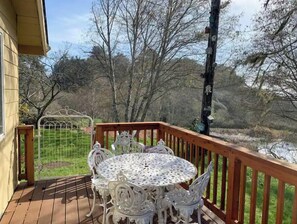 Une terrasse en bois avec une table ronde et des chaises, entourée d’une clôture en métal et d’arbres.