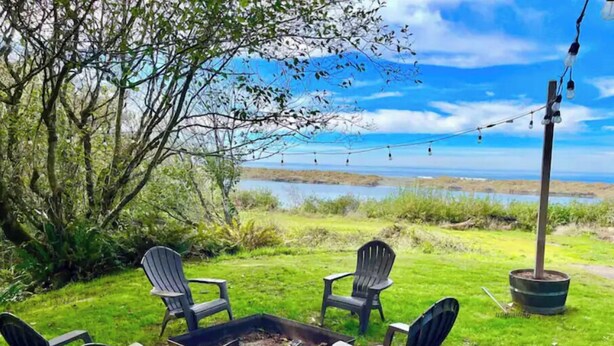 An outdoor seating area with four Adirondack chairs around a fire pit, overlooking a body of water. - Shore Acres Ocean View Cabin and Retreat Space (McKinleyville)
