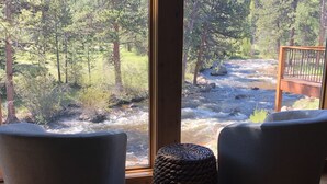 A room with a large window overlooking a flowing river and forested area. There are two modern chairs and a woven basket on the floor. - River Front/.5 mi to EP/ AC/fishing (Estes Park)