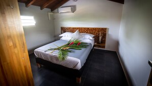 Ein ordentlich bezogenes Bett mit weißen Leinen, einem tropisch gestalteten Mittelstück, einem Holzkopfteil und einer modernen Klimaanlage an der Wand.