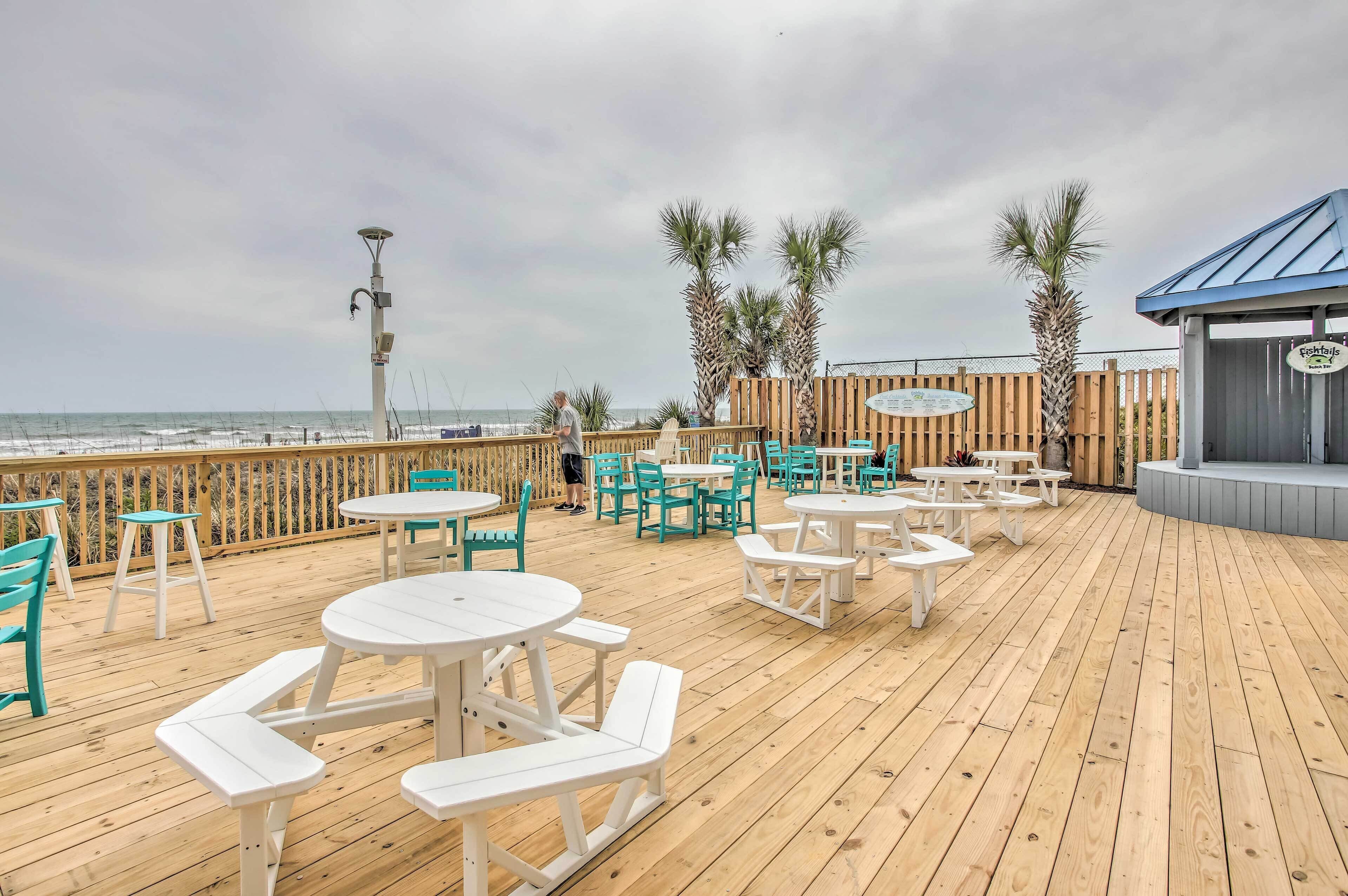 A wooden deck with white tables and chairs, a wooden fence, and a view of the ocean.