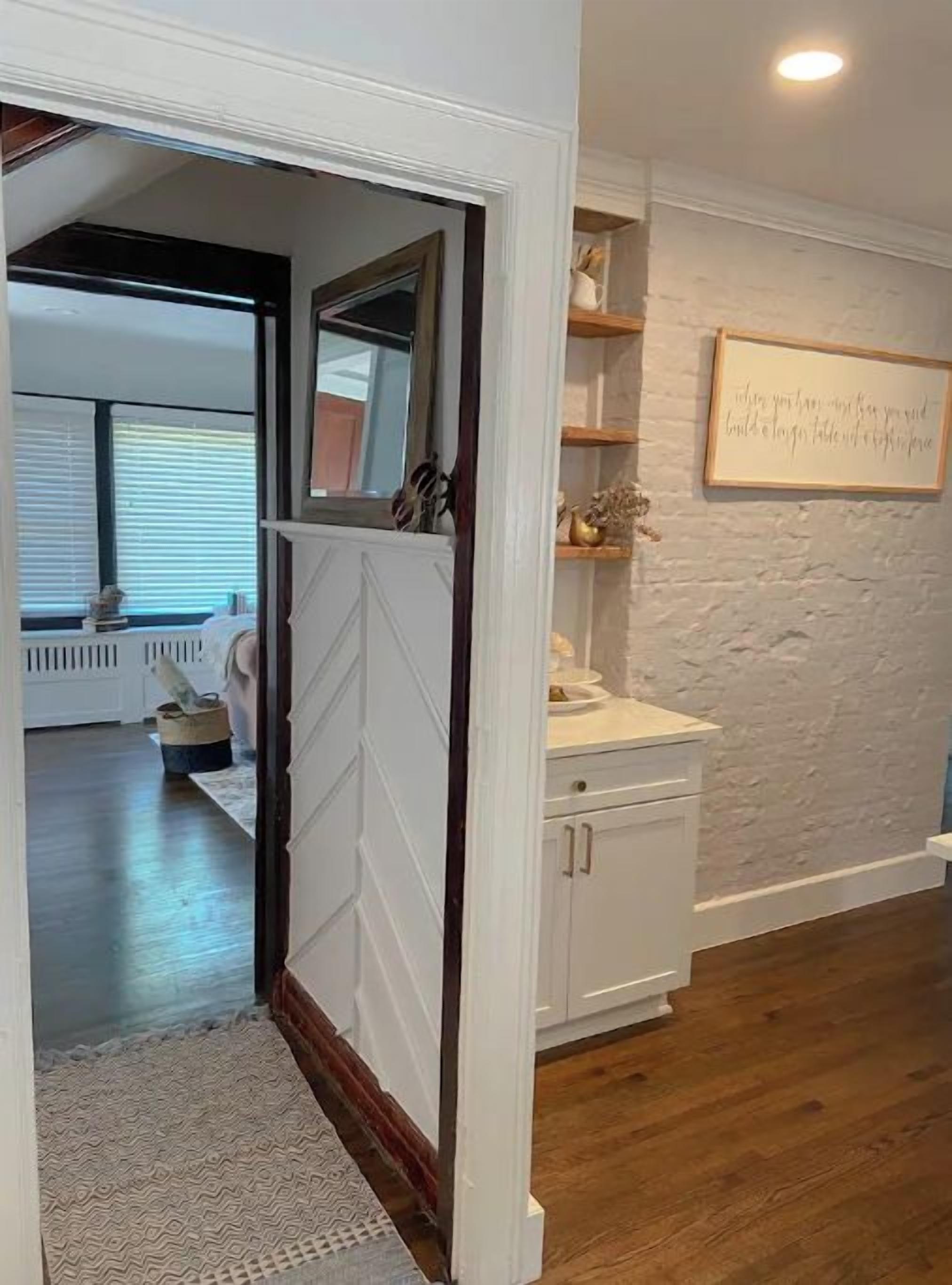 Un couloir doté d’un plancher en bois, de murs blancs et d’une porte blanche donnant sur une autre pièce. Un cadre contenant une citation est accroché au mur, et une petite étagère présente des objets décoratifs.
