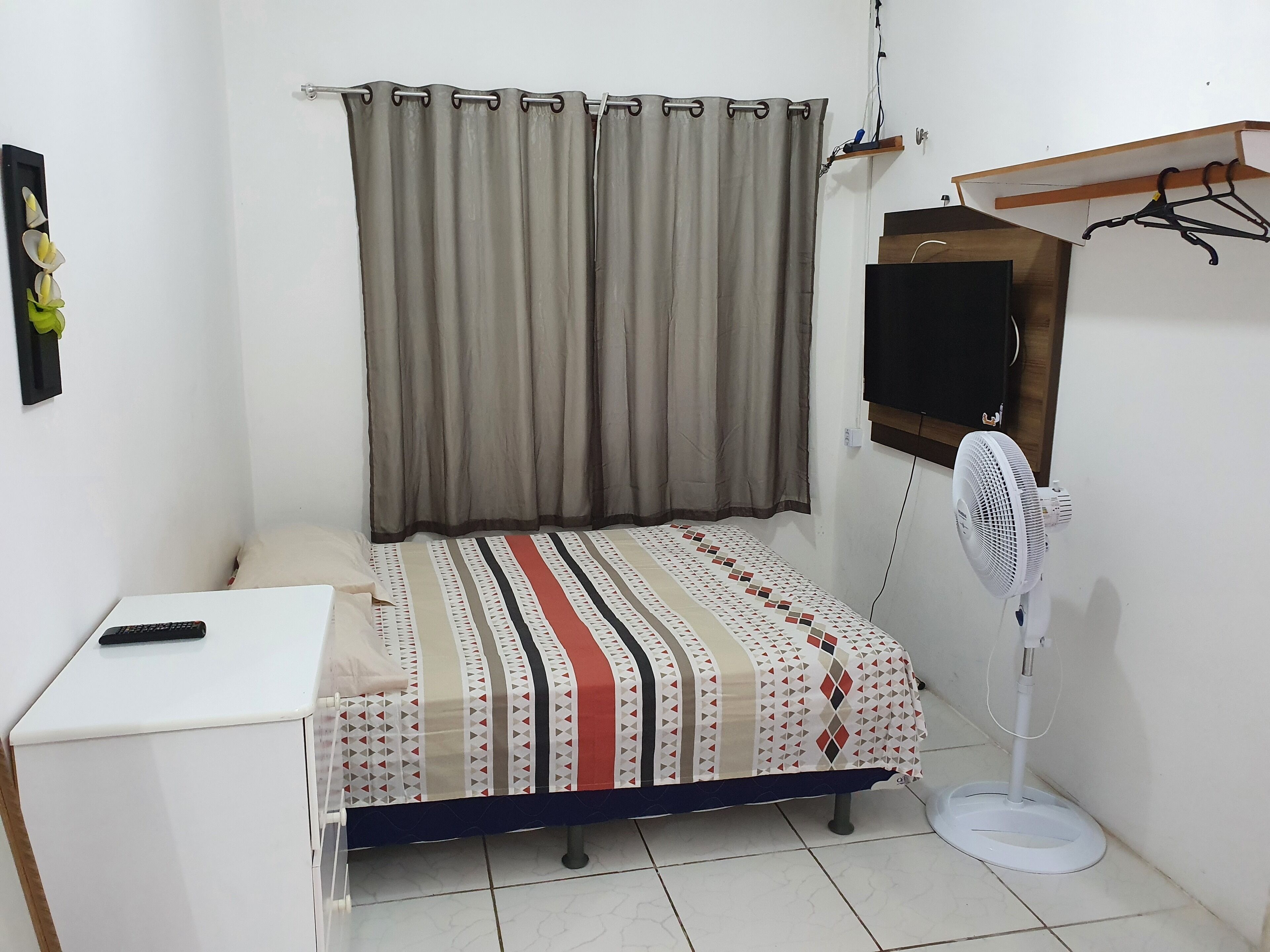 A small, neatly arranged bedroom with a bed, a bedside table, a wall-mounted TV, and a standing fan.