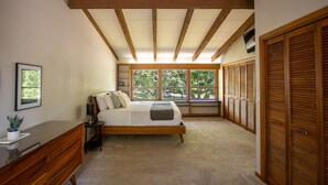 Une chambre à coucher avec un lit en bois, une table de chevet, une commode, une fenêtre donnant sur des arbres et un ensemble d’armoires en bois.