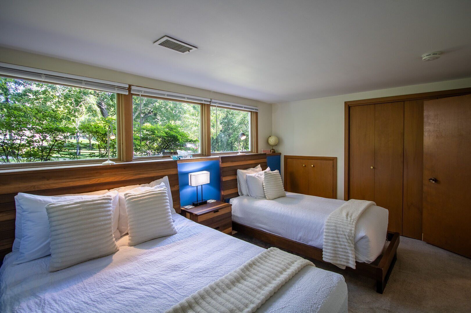 A hotel room with two beds, a wooden headboard, a nightstand with a lamp, and a view of greenery through large windows.