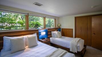 A hotel room with two beds, a wooden headboard, a nightstand with a lamp, and a view of greenery through large windows.