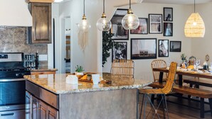 A modern kitchen with a granite countertop, hanging pendant lights, and a dining area with a wooden table and chairs. - Desert Oasis • Hot Tub, Pool, Arcade & Hammocks • Fenced 2 Acres • Joshua Trees (Yucca Valley)