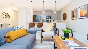 A modern living room with a sofa, coffee table, and chairs. There is a kitchen in the background with cabinets and a refrigerator.