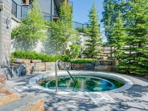 A hot tub surrounded by stone steps and greenery, with a building in the background. - Cozy 2 Bedroom Condo in Silver Creek Lodge (Canmore)