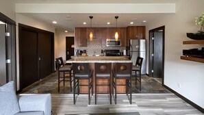 A modern kitchen with a central island, bar stools, and a dining area with a sofa. - Cozy 2 Bedroom Condo in Silver Creek Lodge (Canmore)