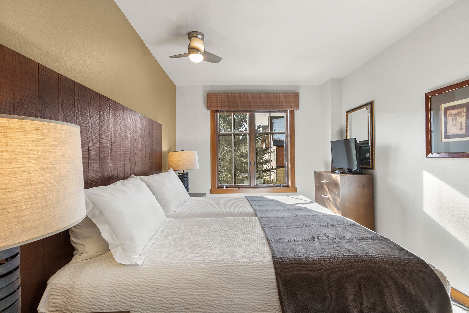 Quarto de hotel com cama, ventoinha no teto, janela com vista, televisão sobre cômoda de madeira e quadro na parede.
