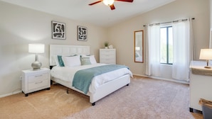 A bedroom with a bed, nightstands, a ceiling fan, a window with curtains, and artwork on the walls. - Hike, Bike, Explore! Mtn-View Tucson Home (Tucson)