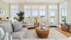 Ein geräumiges Wohnzimmer mit grauem Sofa, gestreiftem Ohrensessel, Couchtisch und großen Fenstern mit Blick ins Freie.