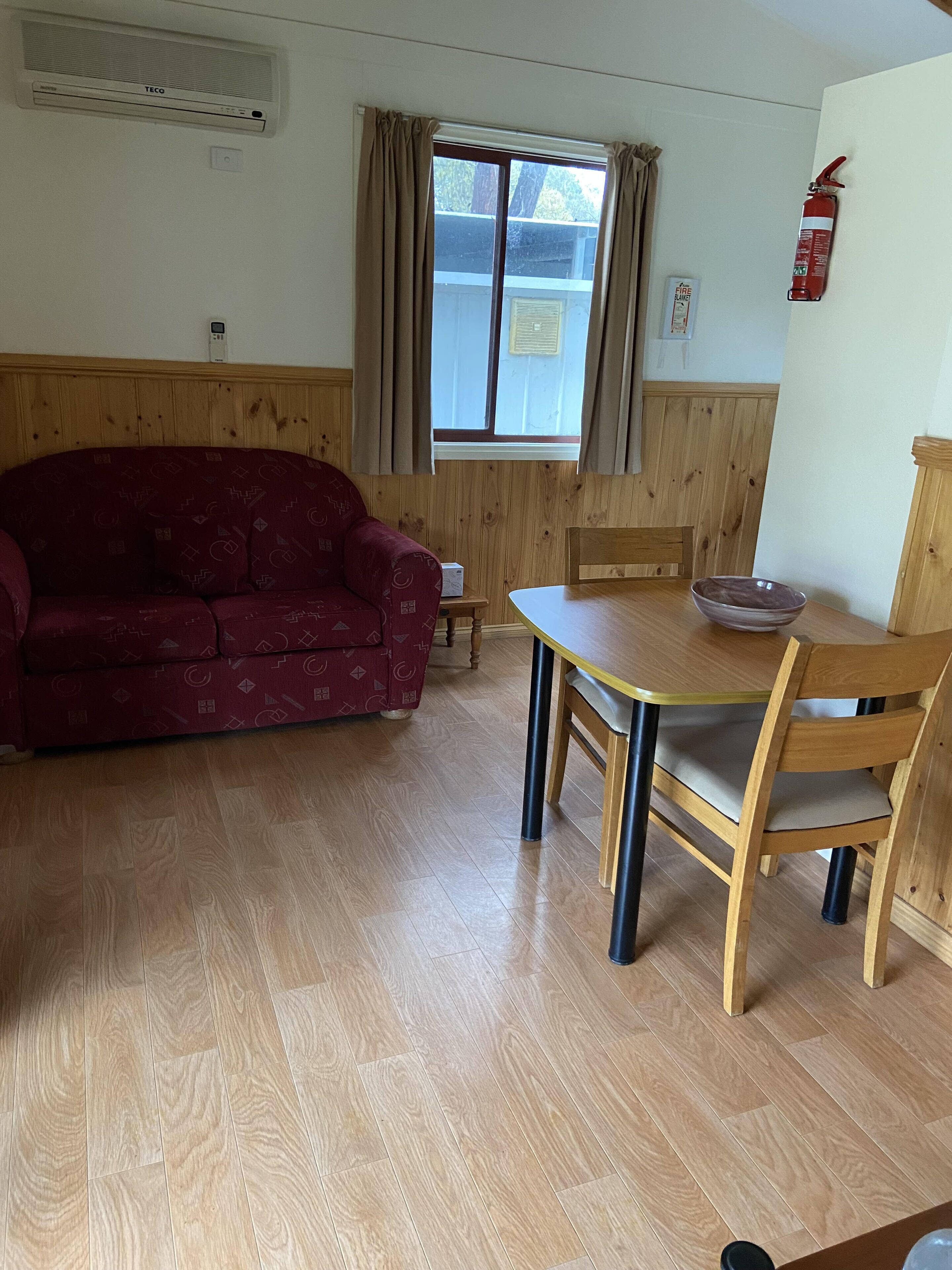 A room with a wooden floor, a red patterned sofa, a wooden dining table with a bowl, and a fire extinguisher on the wall.