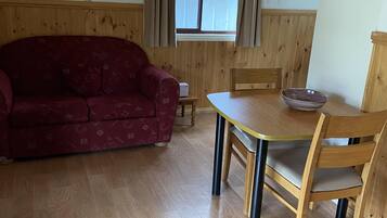 A room with a wooden floor, a red patterned sofa, a wooden dining table with a bowl, and a fire extinguisher on the wall.
