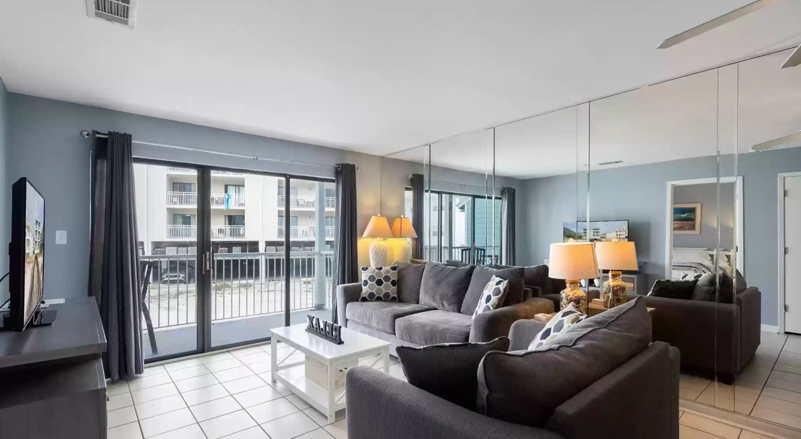 A modern living room with two sofas, a coffee table, and a TV. There are two lamps on the floor. The room has large windows and a balcony.