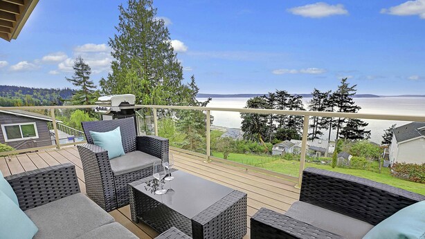 Eine Terrasse mit Korbmöbeln und einem Grill, mit Blick auf einen See und bewaldete Hügel.
