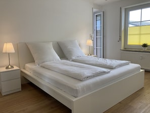 Une chambre à coucher au style minimaliste, avec un lit blanc, deux tables de chevet et une fenêtre équipée de stores.