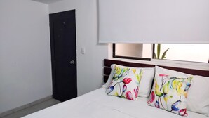 Un dormitorio con cama, una puerta oscura, una ventana con vistas y almohadones florales.