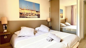 Hotelkamer met een bed, nachtkastje, lamp, schilderij van een haven en een spiegel die de kamer weerspiegelt.