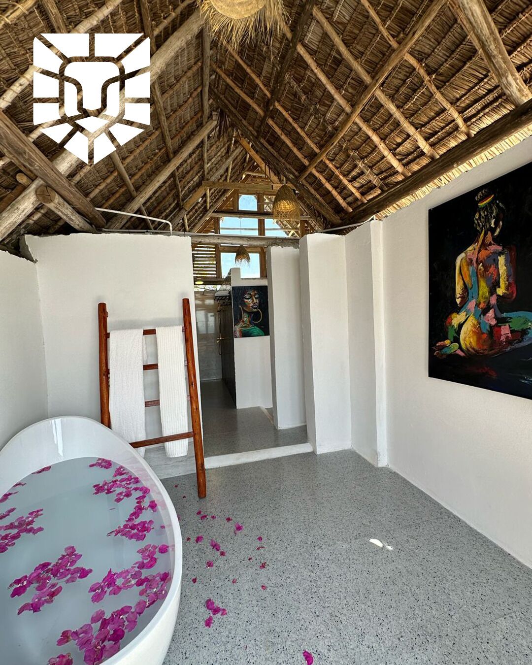 A room with a thatched roof, a bathtub with rose petals, a wooden ladder, and a painting on the wall.