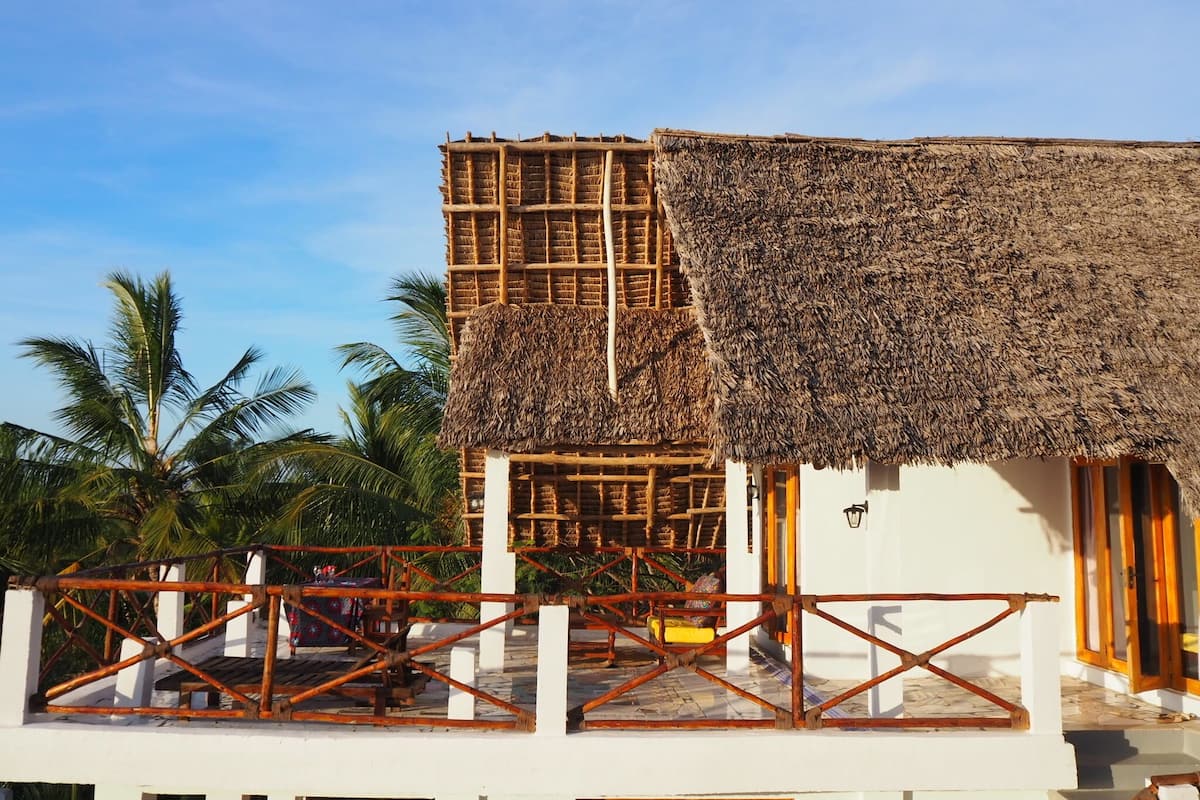 A thatched-roof building with a white exterior, a wooden fence, and palm trees in the background.