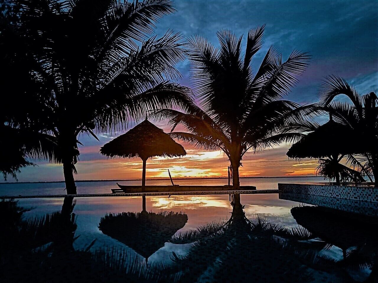 A serene sunset view with palm trees and a thatched umbrella by a reflective pool.