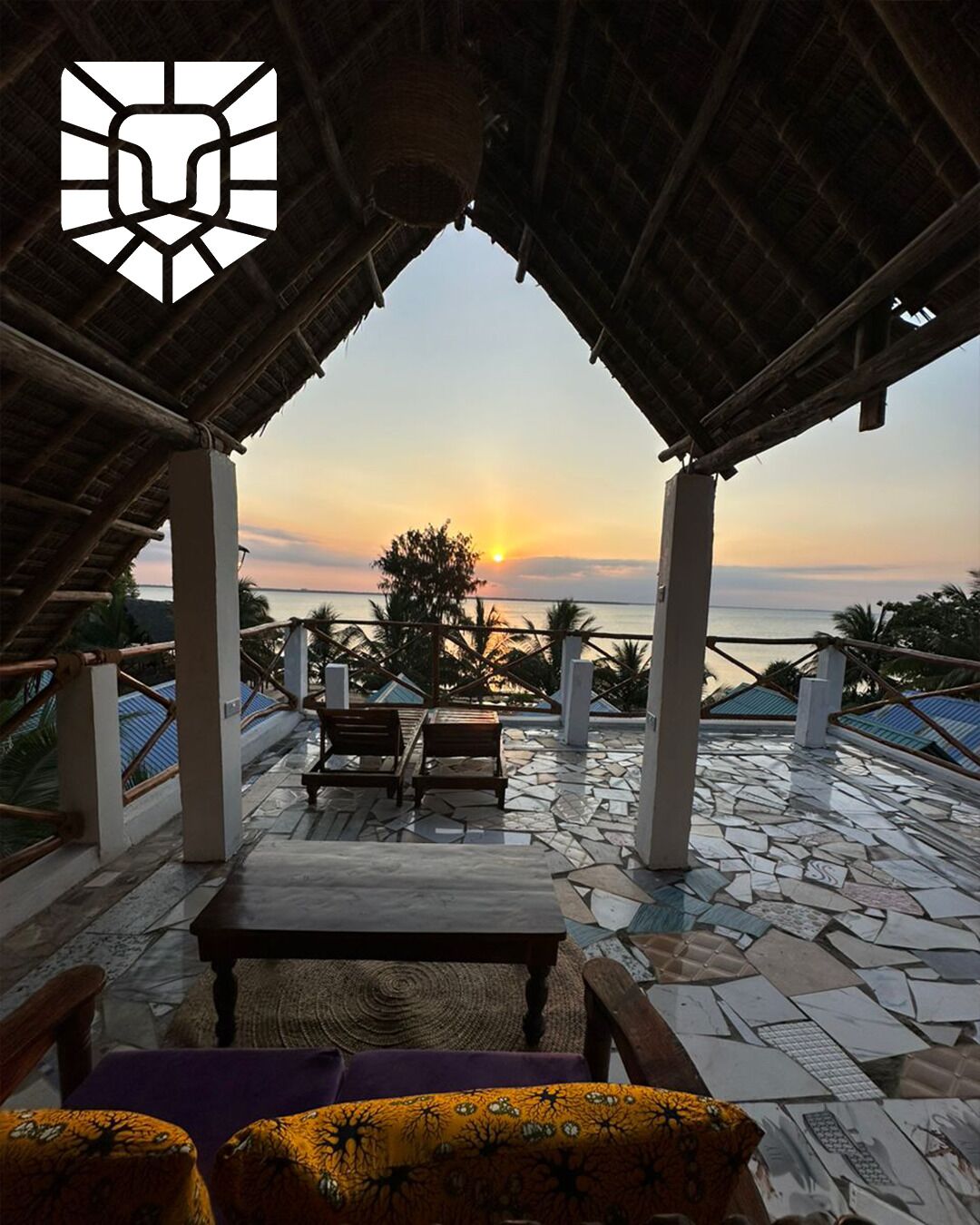 A covered outdoor seating area with a view of the sunset over the sea, stone flooring, and a thatched roof structure.