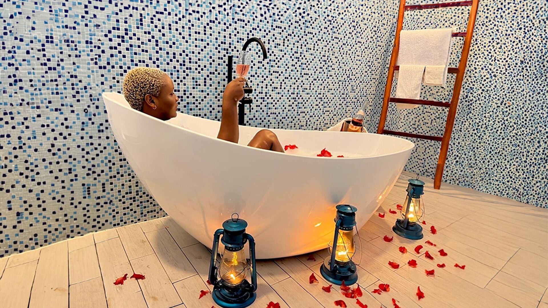 A person relaxing in a large bathtub with a mosaic tile wall in the background.