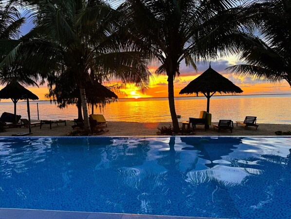A poolside view with a sunset over the ocean, palm trees, and lounge chairs. - Asante Sana Beachfront Hotel (Fukuchani)
