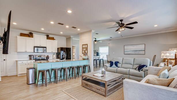 A modern kitchen with white cabinets, a central island with stools, and a dining area with a gray sofa and a ceiling fan. - Savings Alert! Pet-Friendly: Spacious, Fenced Retreat, Views! Games!  Sleeps 14 (Crystal Beach)