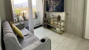 Ett modernt vardagsrum med en grå soffa, en takfläkt, en tv och en balkong med utsikt över byggnader och berg.