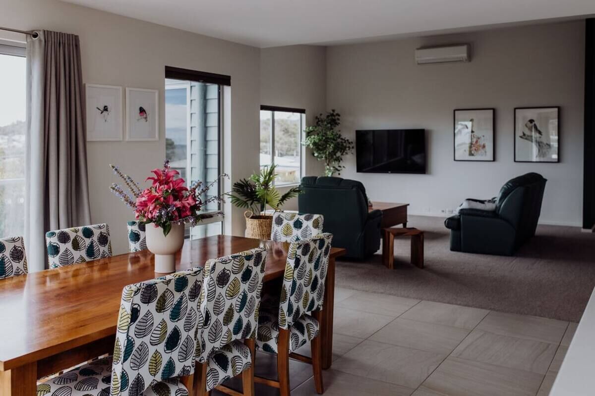 A modern living room with a dining area, featuring a wooden dining table, patterned chairs, a green sofa, and a flat-screen TV on the wall.