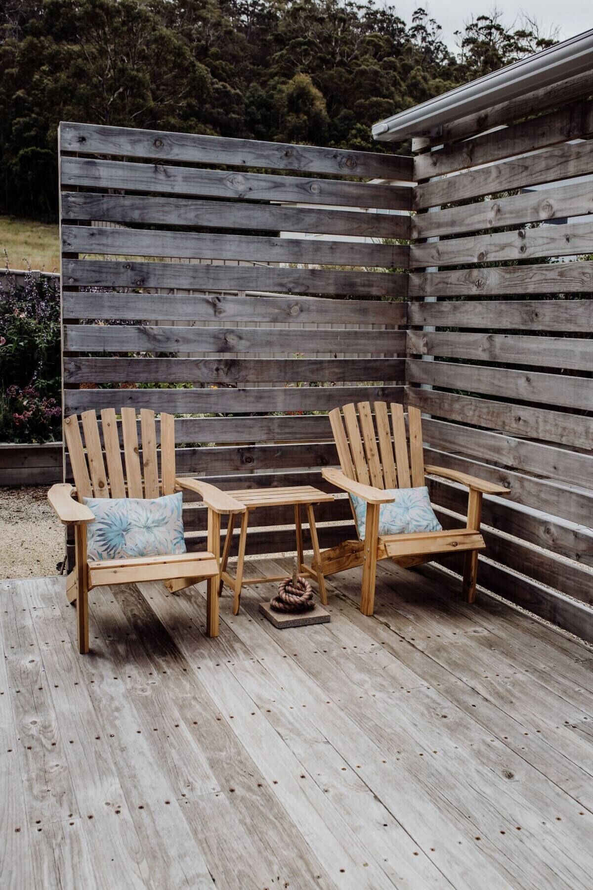 Two wooden Adirondack chairs with cushions on a wooden deck, positioned in front of a wooden fence.