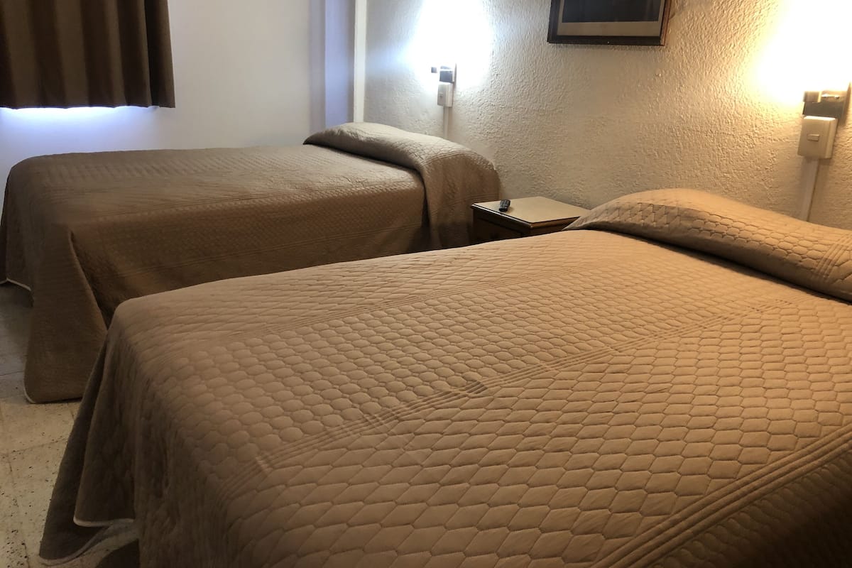 Quarto de hotel com duas camas cobertas por colchas marrons, uma mesa de cabeceira com um controle remoto e uma luminária fixada na parede.