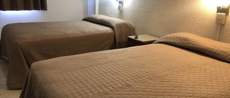 A hotel room with two beds covered in brown quilts, a nightstand with a remote control, and a wall-mounted light.