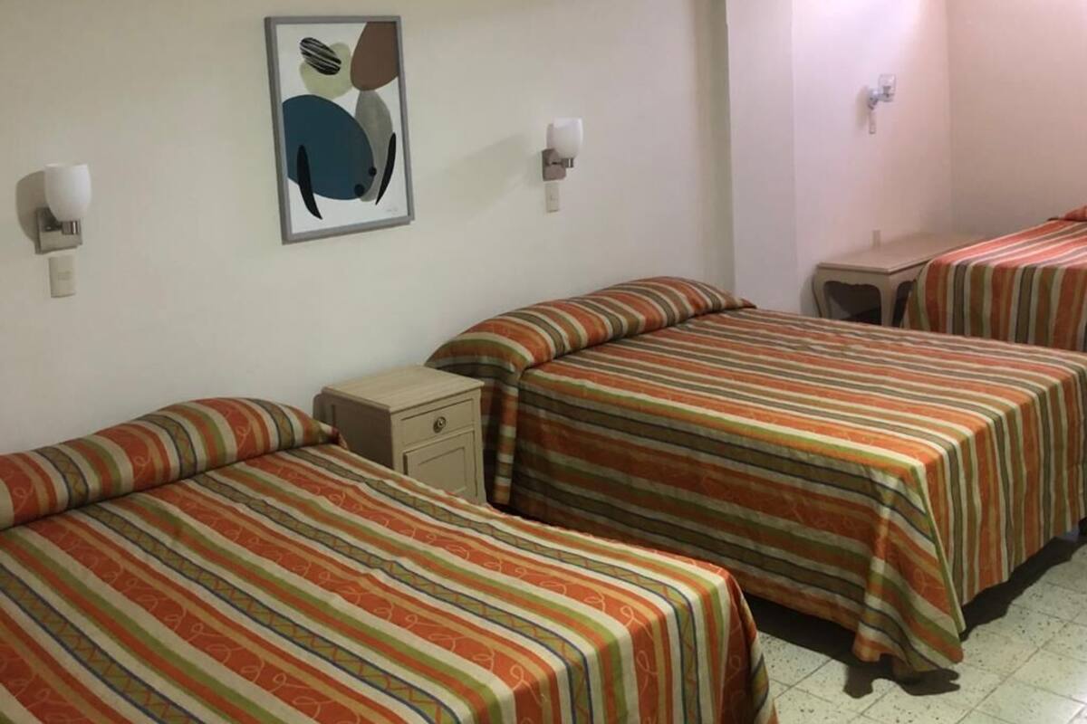 Quarto de hotel com três camas, cada uma com roupa de cama listrada, uma pequena mesa de cabeceira, uma luminária na parede e um quadro emoldurado.